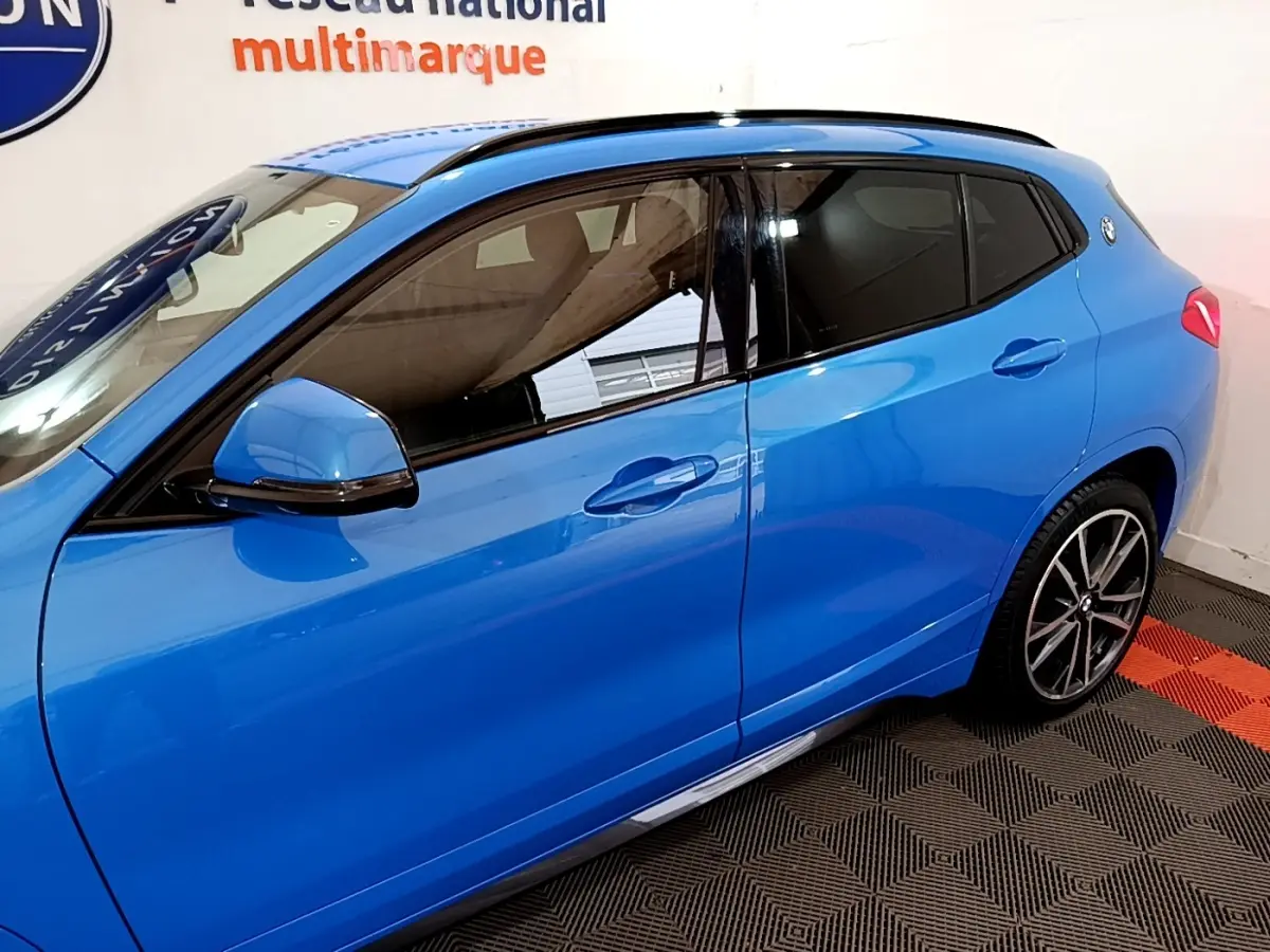 Vue côté gauche d'un BMW X2 sDrive20i M Sport bleu, avec jantes alliage et vitres teintées en intérieur showroom.