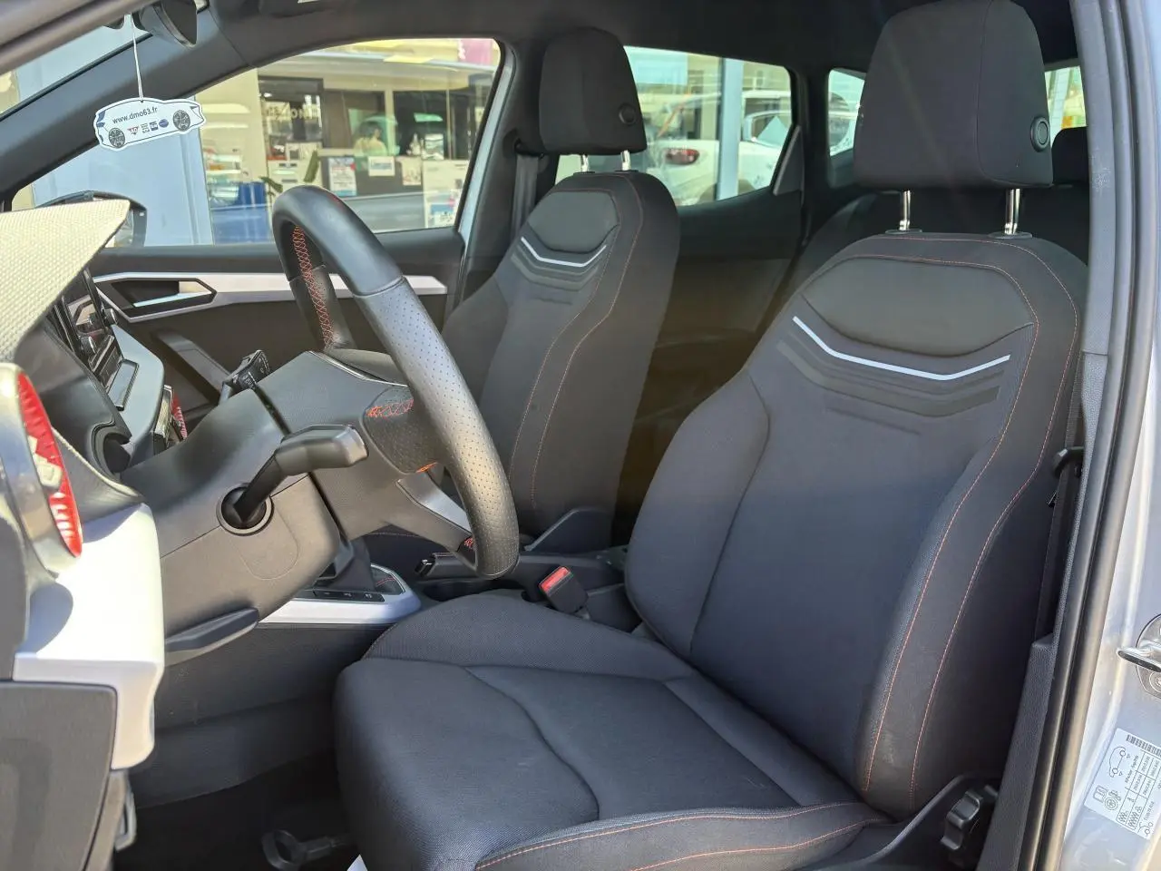 Vue intérieure côté conducteur du SEAT ARONA 2024, sièges textile noirs avec surpiqûres rouges et volant sport cuir.