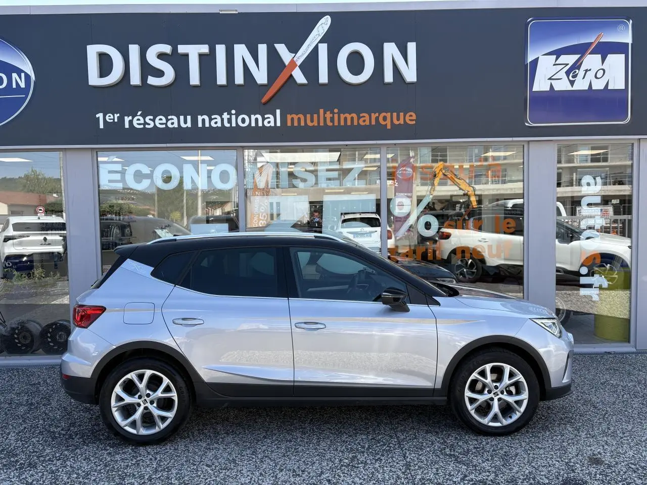 Profil latéral droit d'une SEAT Arona 2024 GRS avec toit noir et jantes alliage 16 pouces devant une vitrine Distinxion.