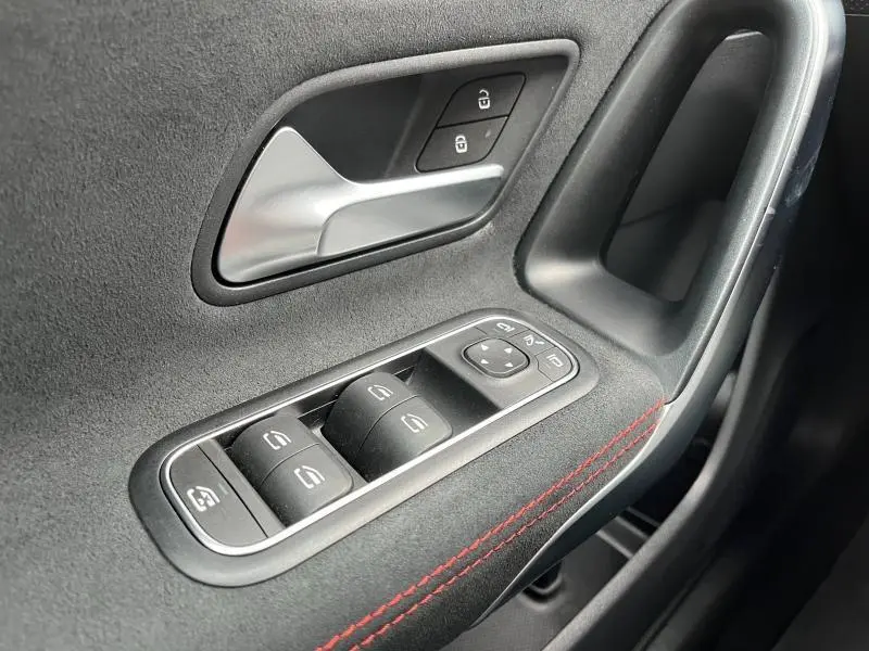 Gros plan sur la commande des vitres et la poignée intérieure côté gauche en noir avec surpiqûres rouges dans une Mercedes CLA Shooting Brake.