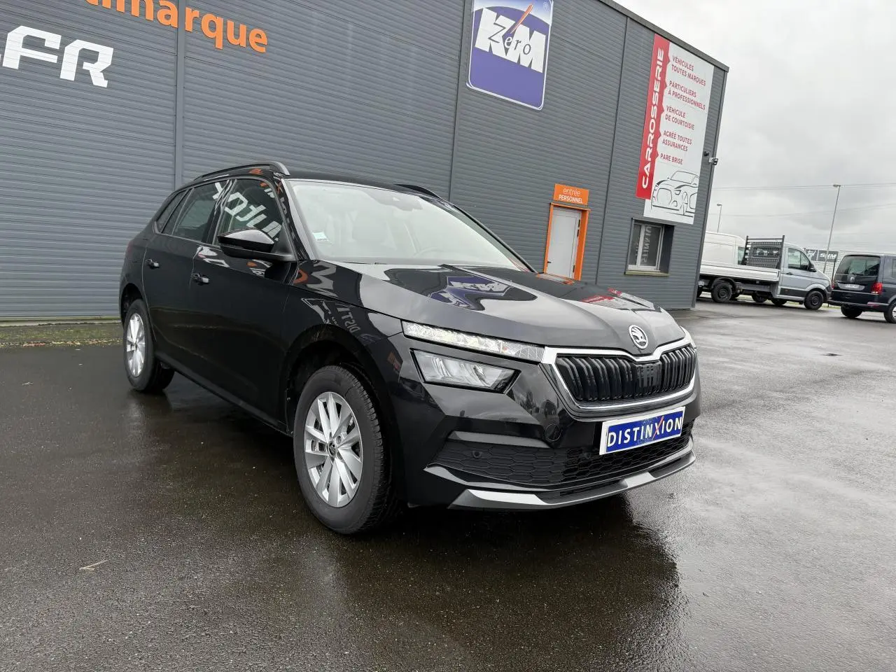 Vue 3/4 avant gauche d'un Skoda Kamiq noir magic nacré avec calandre chromée, stationné devant un bâtiment gris.