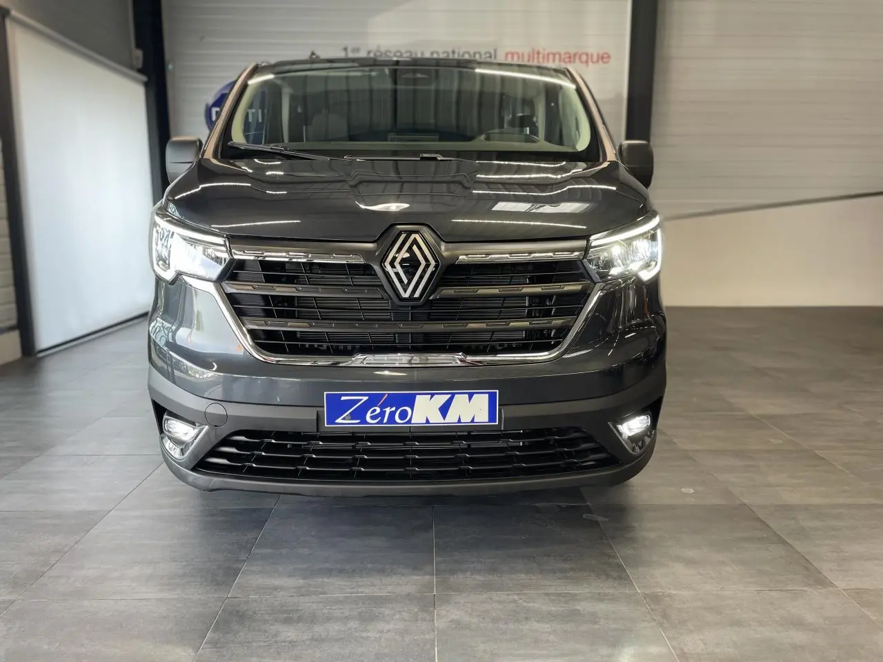 Vue frontale du Renault Trafic Fourgon L2H1 2025 en gris comète avec phares et feux LED allumés dans un showroom.