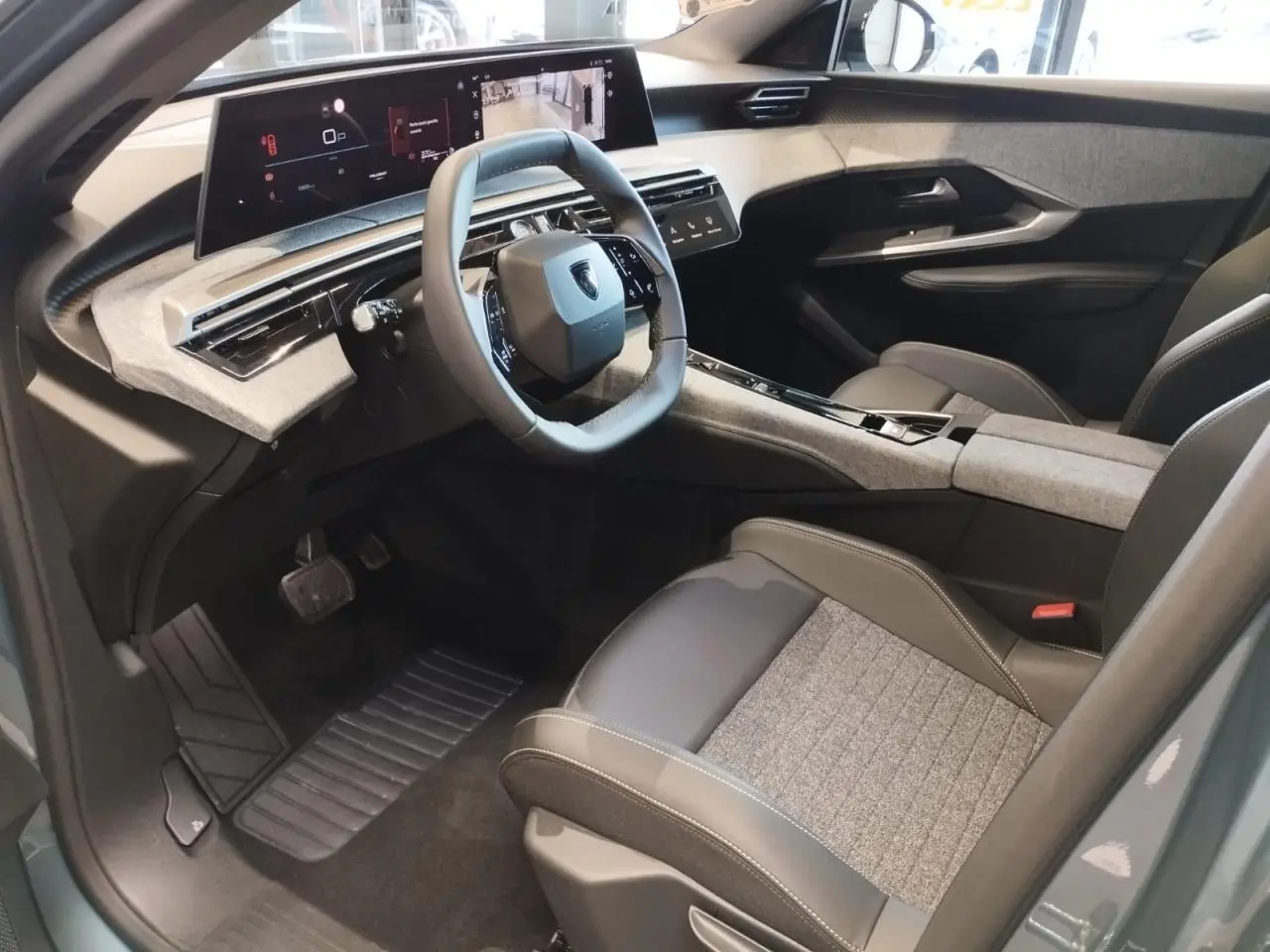 Vue intérieure côté conducteur du Peugeot 3008 Hybrid 2025 avec tableau de bord et écran incurvé, sellerie tissu gris et cuir noir.
