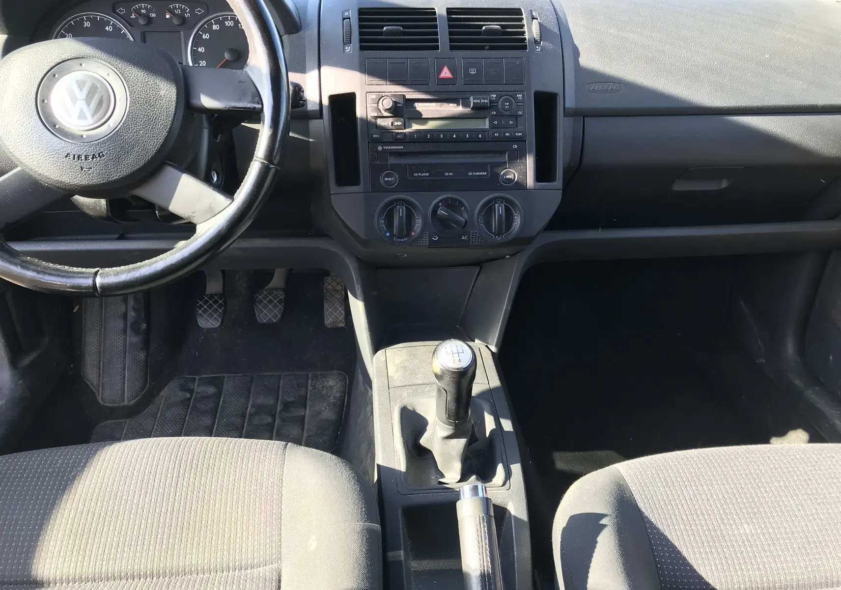 Intérieur avant de la Volkswagen Polo Match 2004, vue centrée sur le tableau de bord, volant, levier de vitesse manuel et sièges tissu noir.