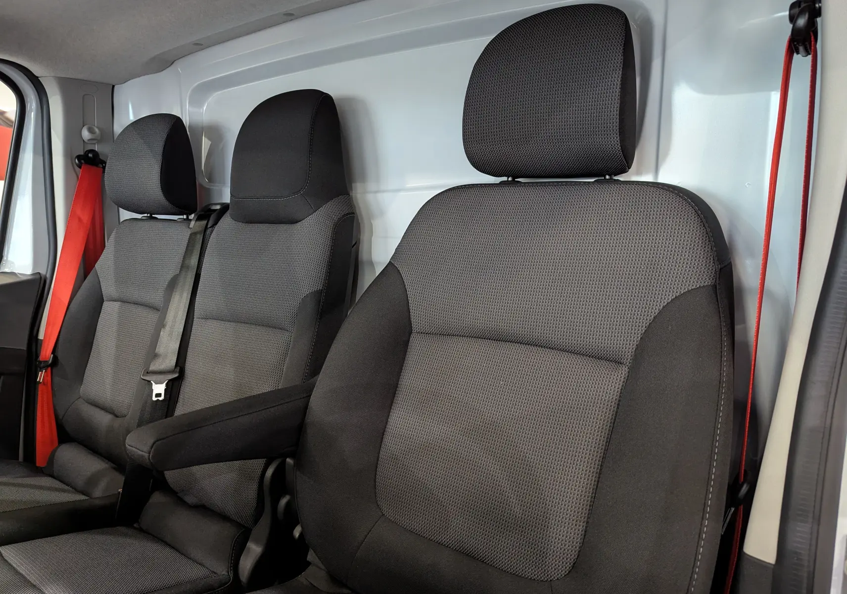 Vue intérieure des sièges avant noirs en tissu du Renault Trafic Fourgon blanc, avec ceinture rouge visible côté gauche.