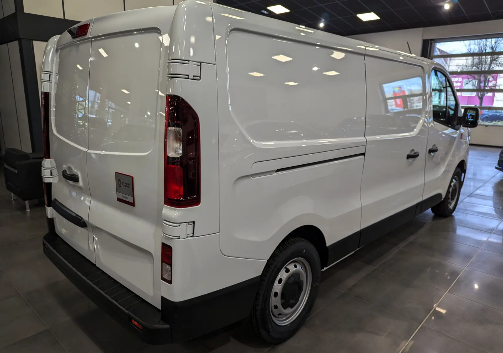 Vue 3/4 arrière droite d'un Renault Trafic Fourgon blanc minéral 2025 dans un showroom lumineux.