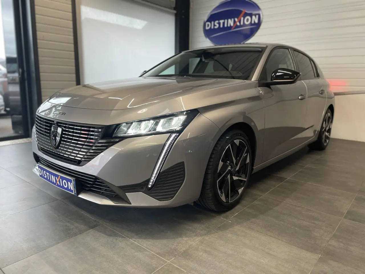 Peugeot 308 gris Artense vue 3/4 avant droit en showroom avec calandre noire et feux LED distinctifs.