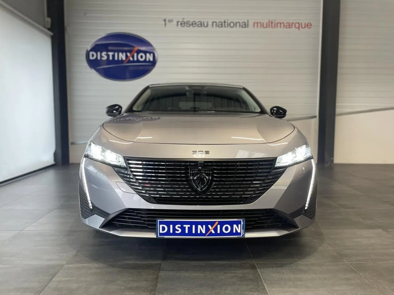 Vue frontale d'une Peugeot 308 gris Artense avec calandre noire et feux LED allumés en intérieur showroom.