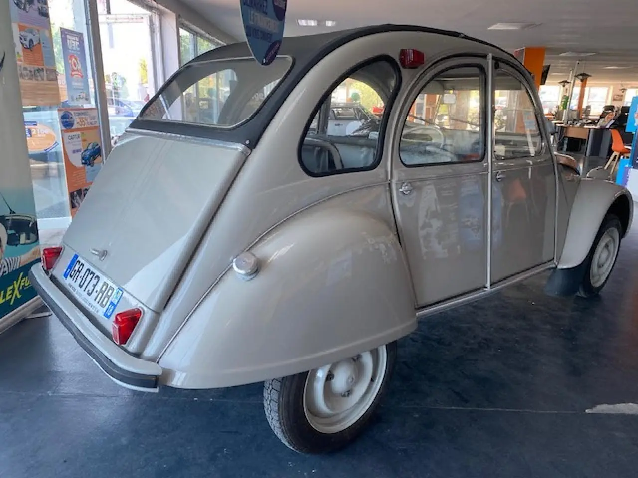 Vue 3/4 arrière droite d'une Citroën 2 CV beige clair refaite à neuf, exposée en intérieur avec ses phares arrière rouges.