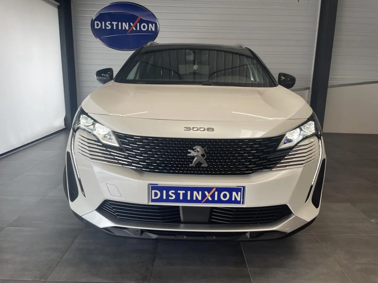 Vue frontale d'un Peugeot 3008 blanc nacré avec calandre noire et phares LED allumés en intérieur showroom.