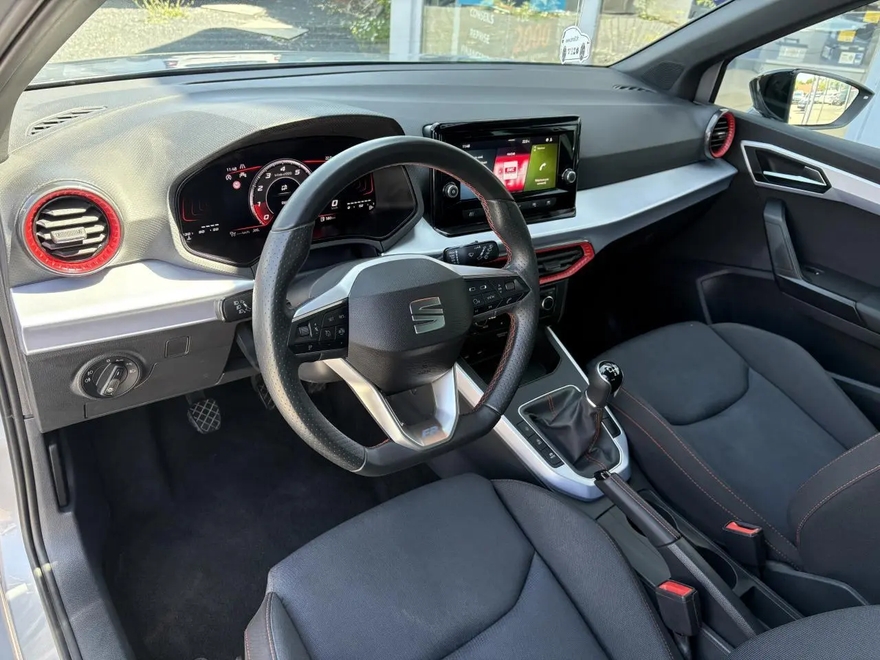 Vue intérieure côté conducteur du SEAT Arona 2024, volant sport cuir noir avec surpiqûres rouges et tableau de bord digital.