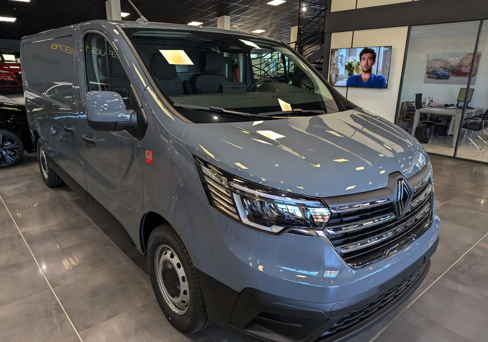 Renault Trafic Fourgon gris urbain en vue 3/4 avant droit, avec calandre chromée et phares LED en showroom.