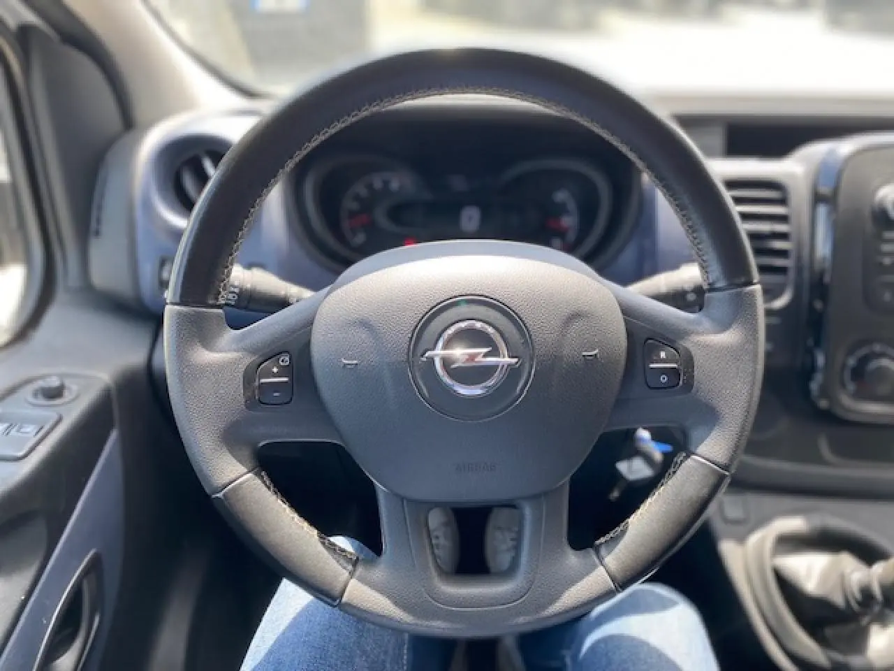 Vue rapprochée du volant noir de l'Opel Vivaro 2018 avec commandes intégrées et tableau de bord flou en arrière-plan.