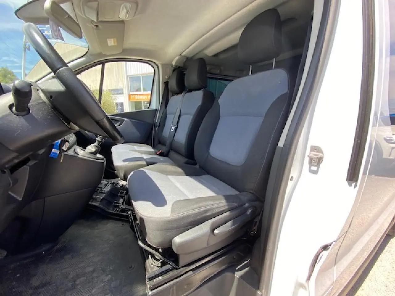 Vue intérieure côté conducteur de l'Opel Vivaro blanc 2018, montrant les sièges avant en tissu gris et noir.