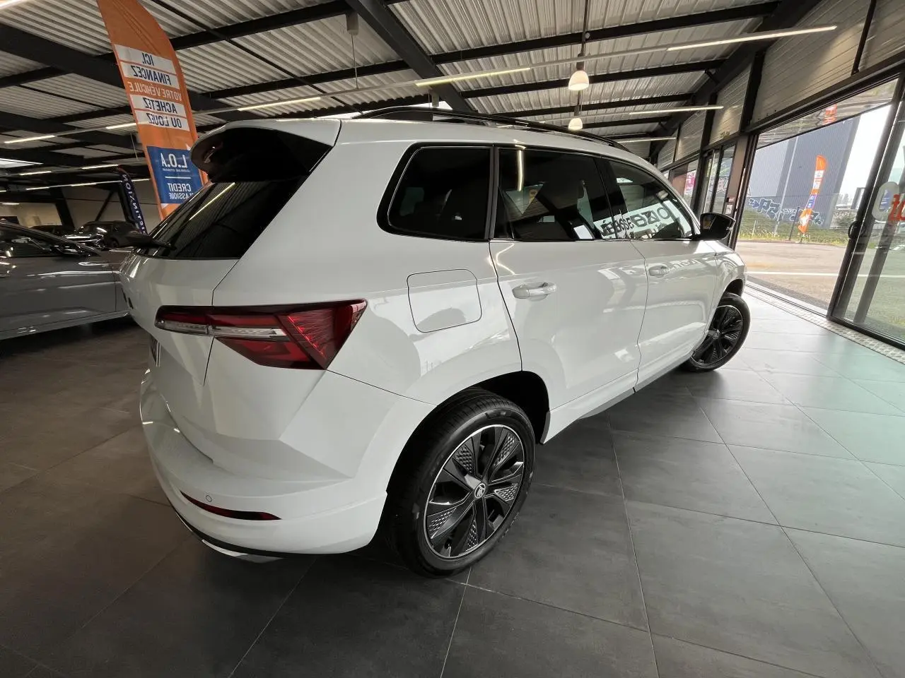 Vue 3/4 arrière droite d'un SUV Skoda Karoq blanc 2025 avec jantes alliage noires et vitres surteintées en showroom.