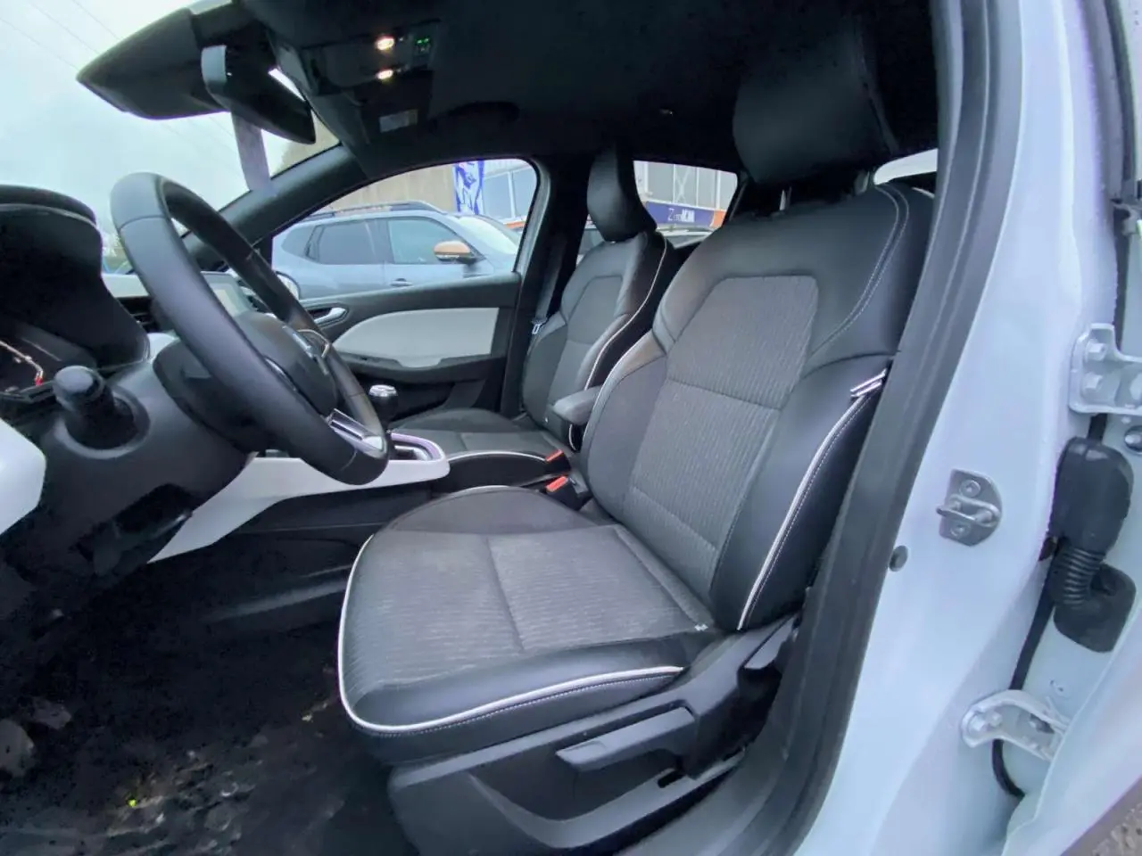 Vue intérieure côté conducteur d'une Renault Clio blanche 2023 avec sièges similicuir gris et tableau de bord moderne.