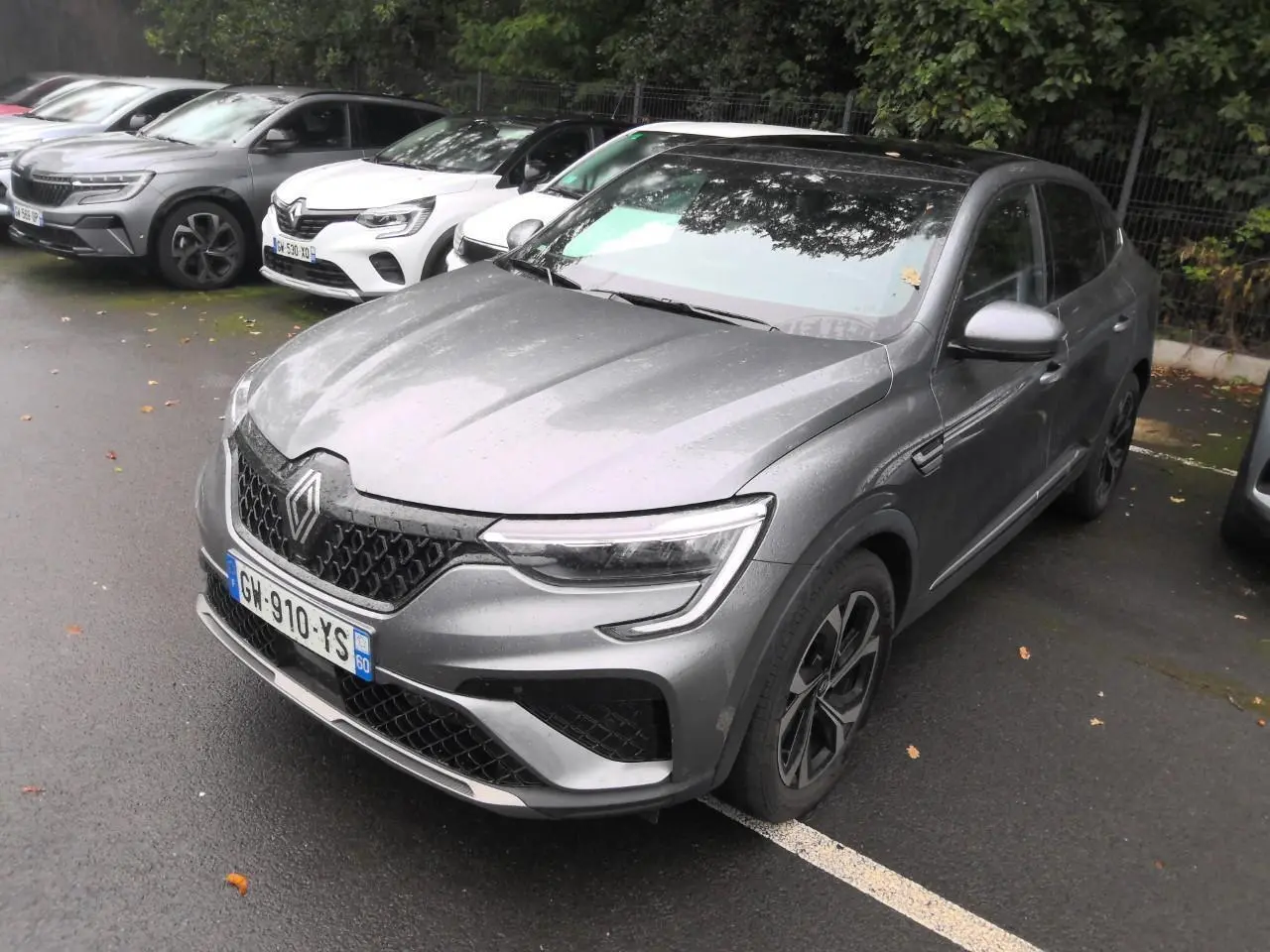 Renault Arkana gris métal foncé vu en 3/4 avant droit sur un parking sous un ciel couvert.