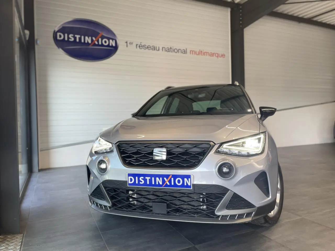 Vue avant d'un SEAT Arona 2024 gris clair avec calandre noire et phares allumés en intérieur showroom.