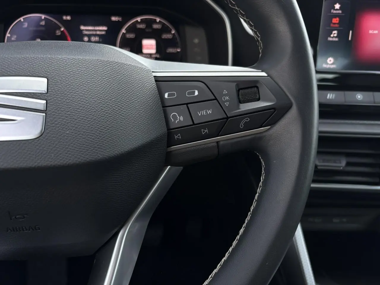 Gros plan sur le volant cuir multifonction de la SEAT LEON 2024 avec commandes intégrées et tableau de bord numérique en arrière-plan.
