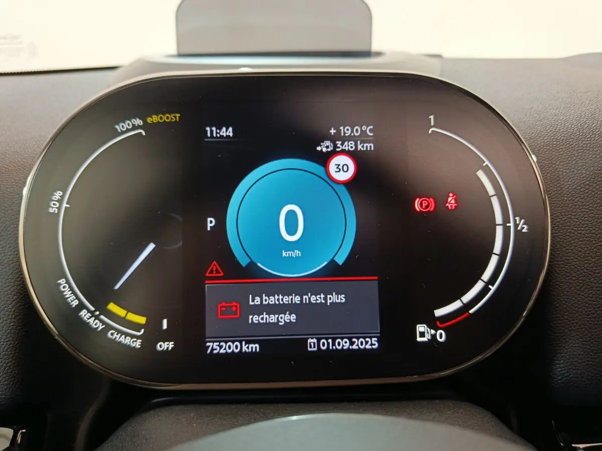 Gros plan sur le tableau de bord numérique du MINI Countryman blanc, affichant la vitesse à 0 km/h et une alerte batterie.