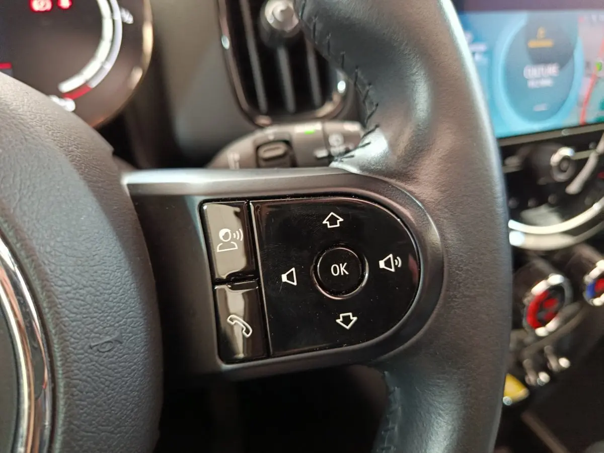 Gros plan sur les commandes multifonctions du volant cuir noir d'un MINI Countryman blanc, avec tableau de bord numérique en arrière-plan.