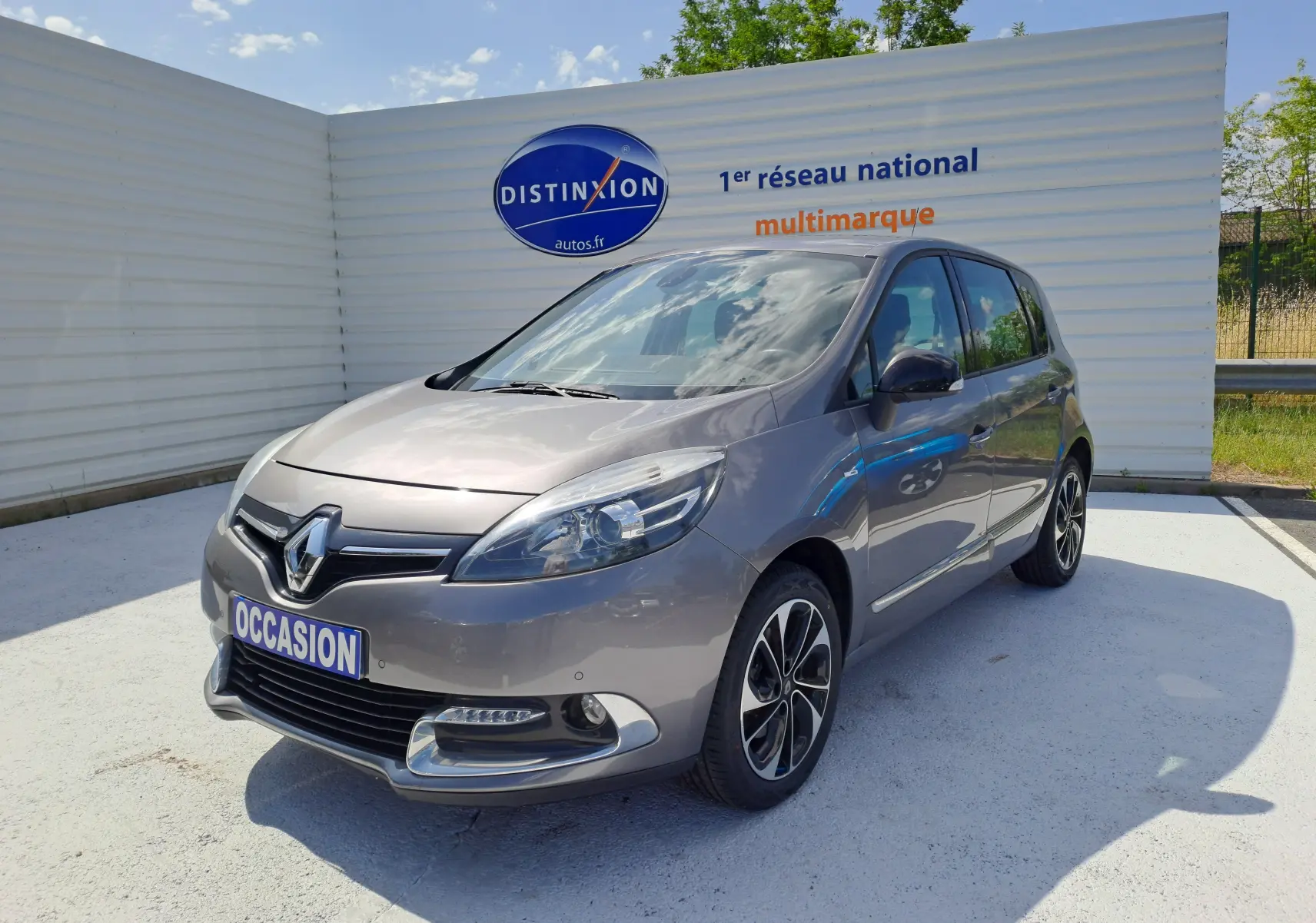 Renault Scenic III gris en 3/4 avant droit, avec jantes alliage noires et logo Renault bien visible sur calandre.