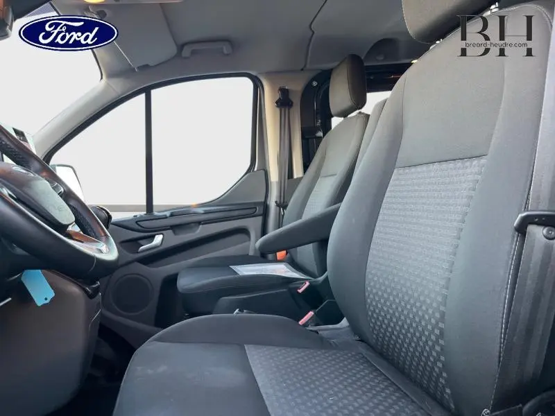 Vue intérieure côté conducteur du Ford Transit Custom Fourgon 2023, sièges gris avec volant cuir et tableau de bord partiel visible.