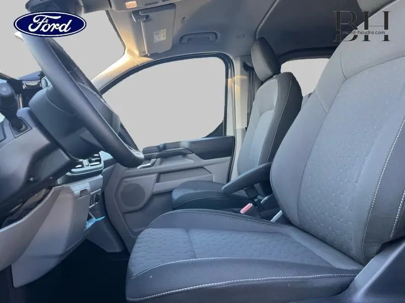 Vue intérieure côté conducteur du Ford Transit Custom Fourgon 2025, sièges gris avec accoudoir et volant visible.