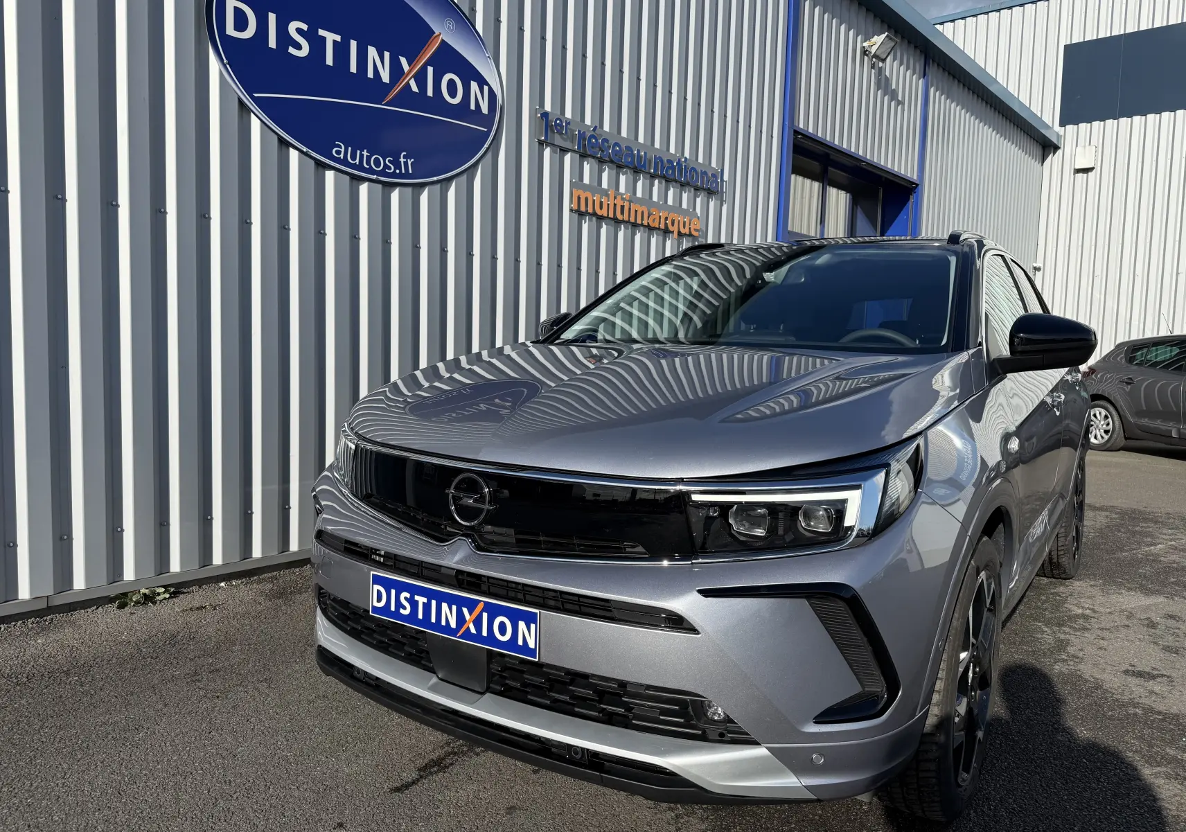 Opel Grandland gris Artense avec toit noir Perla Nera, vue 3/4 avant droit, devant un bâtiment industriel.