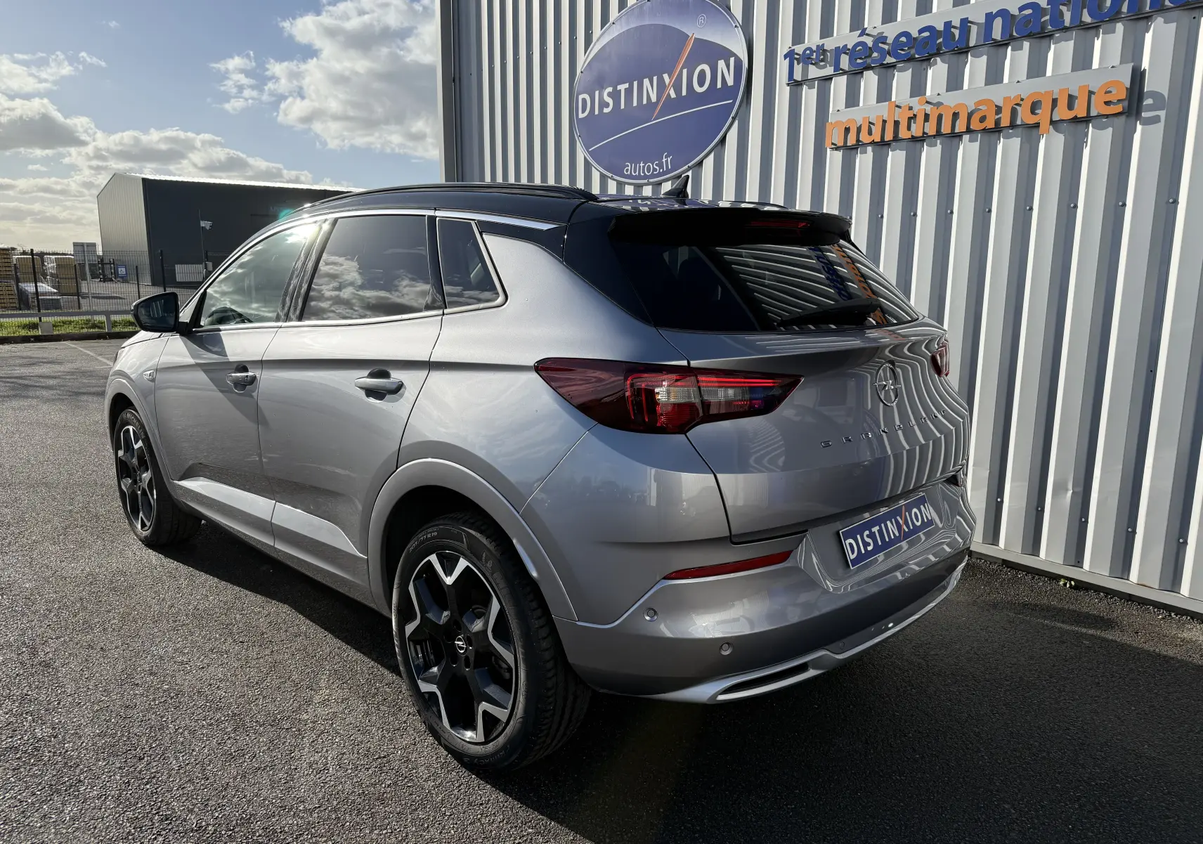 Vue 3/4 arrière droite d'un Opel Grandland gris Artense avec toit noir Perla Nera, garé devant un mur métallique.