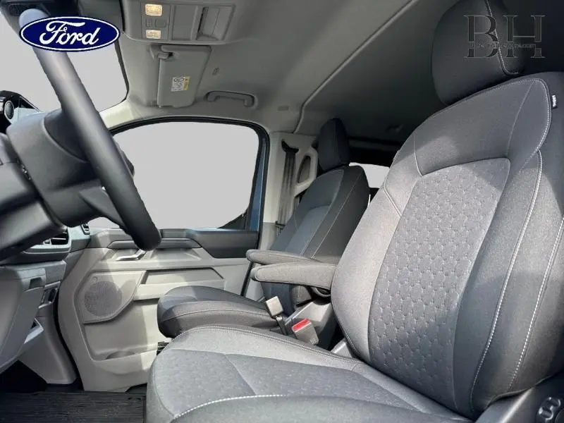Vue intérieure côté conducteur du Ford Transit Custom Fourgon 2025, sièges tissu gris avec accoudoir et détails noirs.