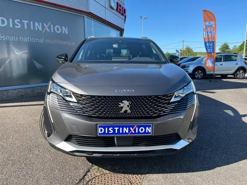 Vue frontale d'un Peugeot 5008 gris Artense 2024 avec calandre noire et logo lion chromé visible au soleil.