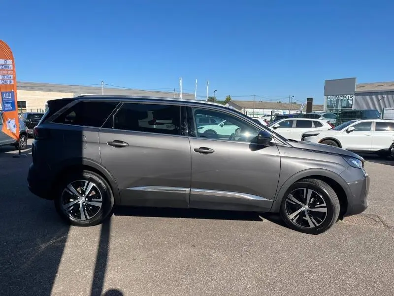 Profil côté droit du SUV Peugeot 5008 gris Artense 2024 avec jantes alliage et barres de toit visibles en extérieur.