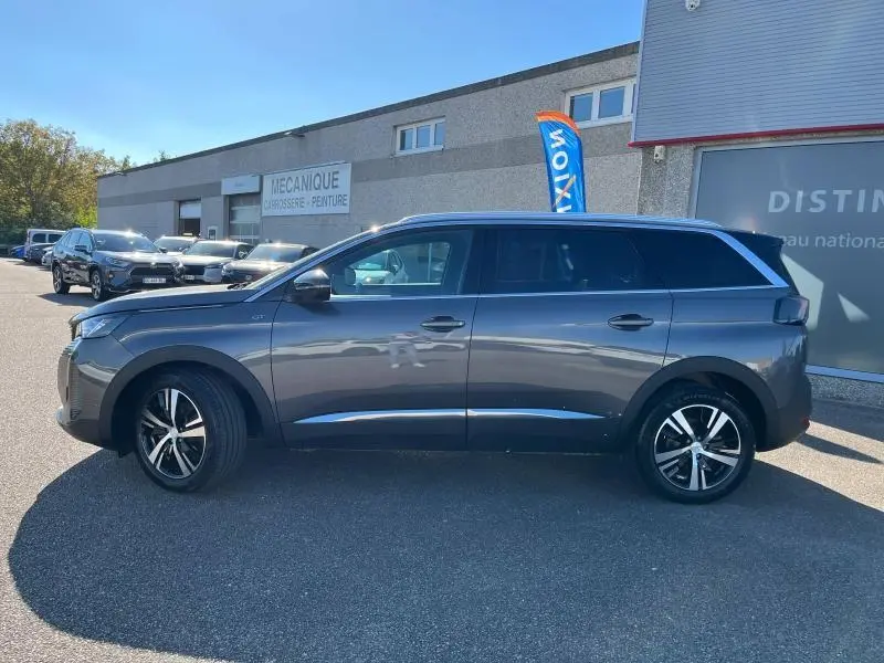 Profil côté gauche du Peugeot 5008 gris Artense 2024, avec jantes alliage et vitres surteintées, stationné devant un atelier mécanique.