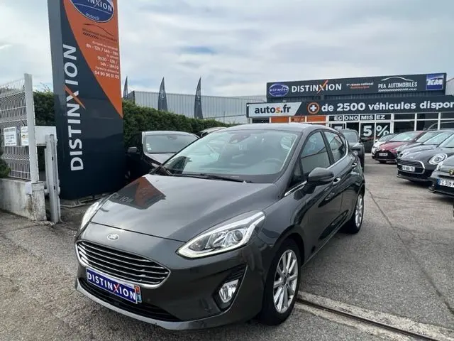 Ford Fiesta 2018 gris vue 3/4 avant droit sur parking avec panneau Distinxion en arrière-plan
