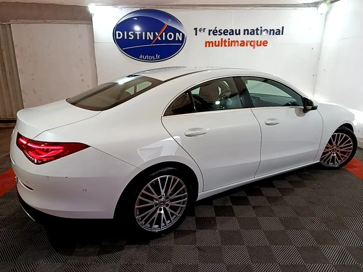 Vue 3/4 arrière droite d'une Mercedes CLA 200 d Business Line blanche avec feux arrière LED allumés dans un showroom.