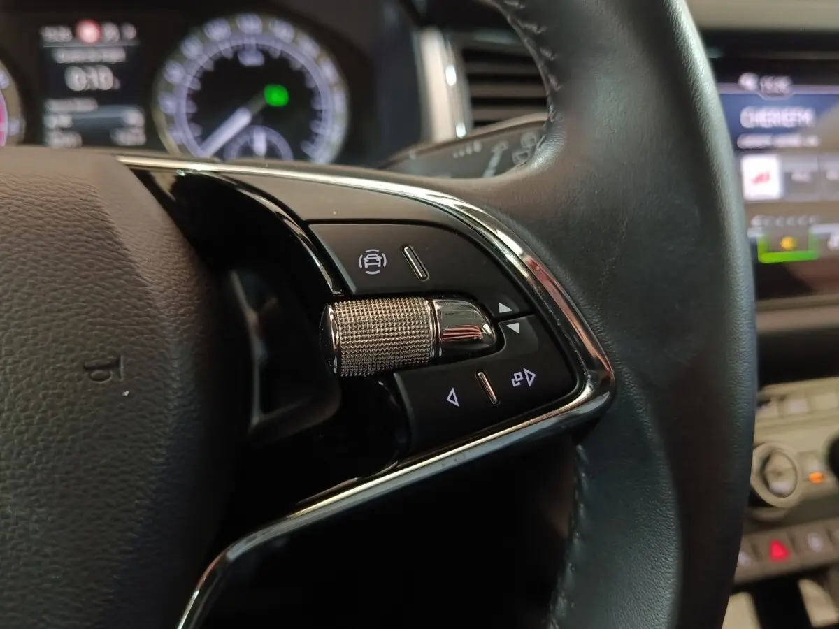 Gros plan sur les commandes multifonctions côté gauche du volant cuir noir d’un Skoda Kodiaq blanc, affichage digital en arrière-plan.