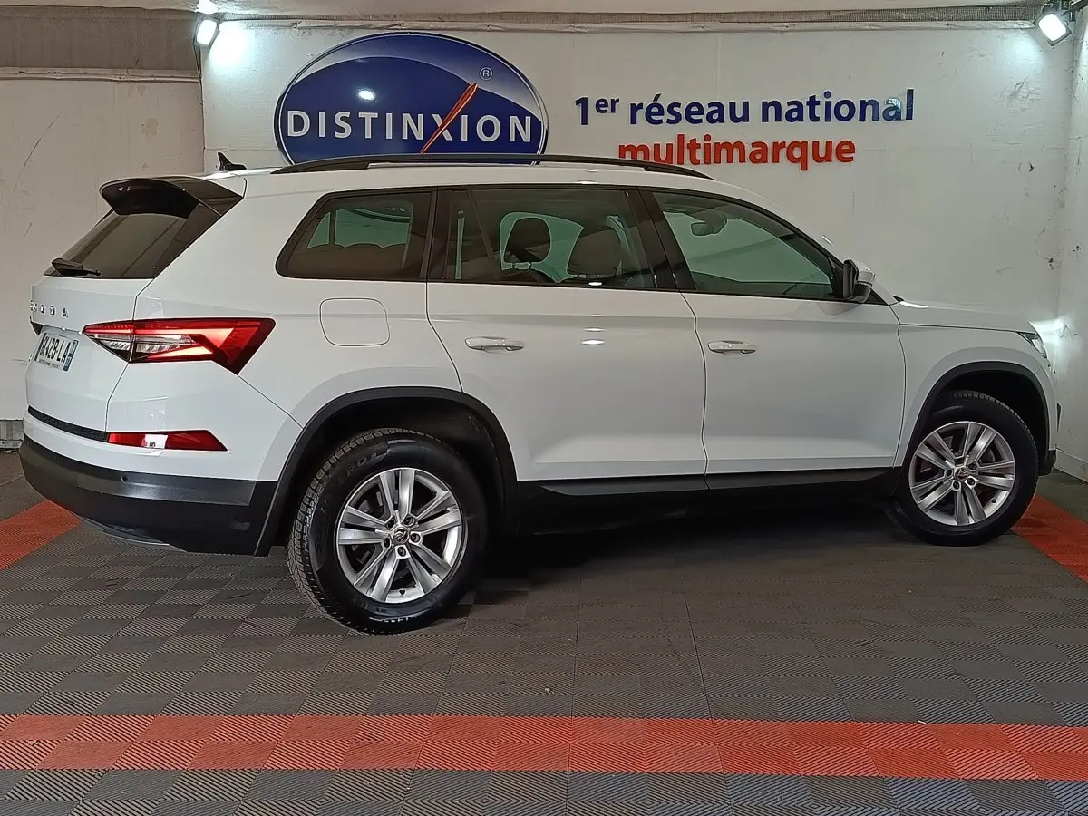 Vue de profil côté gauche d'un Skoda Kodiaq blanc 7 places, avec jantes alliage et toit noir en arrière-plan intérieur showroom.