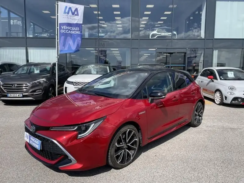 Toyota Corolla 184 H Collection rouge métal avec toit noir, vue 3/4 avant droit, garée devant un showroom.