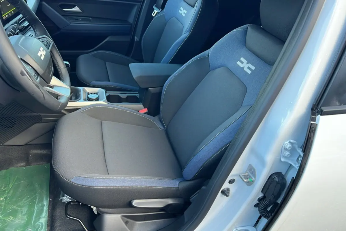 Vue intérieure côté conducteur du Dacia Duster blanc glacier 2025, sièges tissu bleu et noir avec logo distinctif brodé.