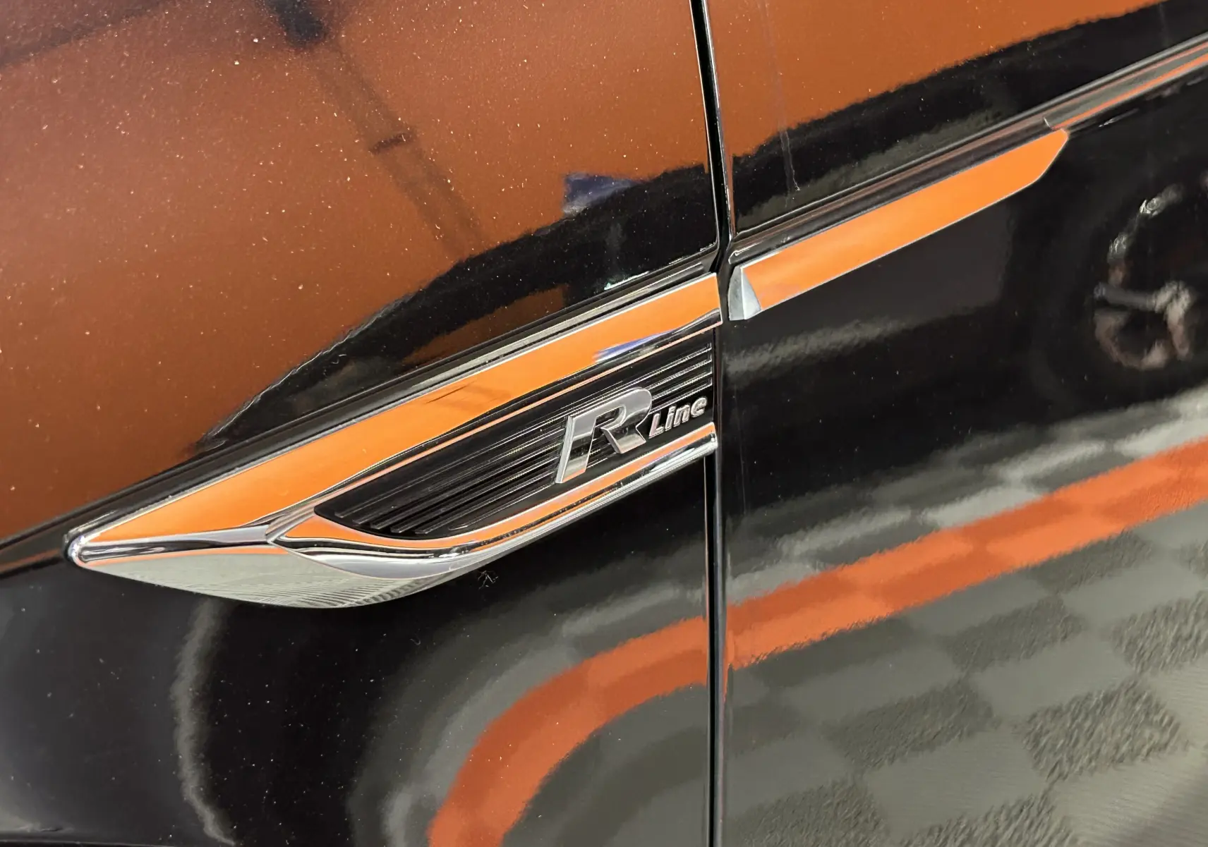Détail du logo R-Line sur la portière noire d'un Volkswagen T-Roc, avec reflet orange au sol.