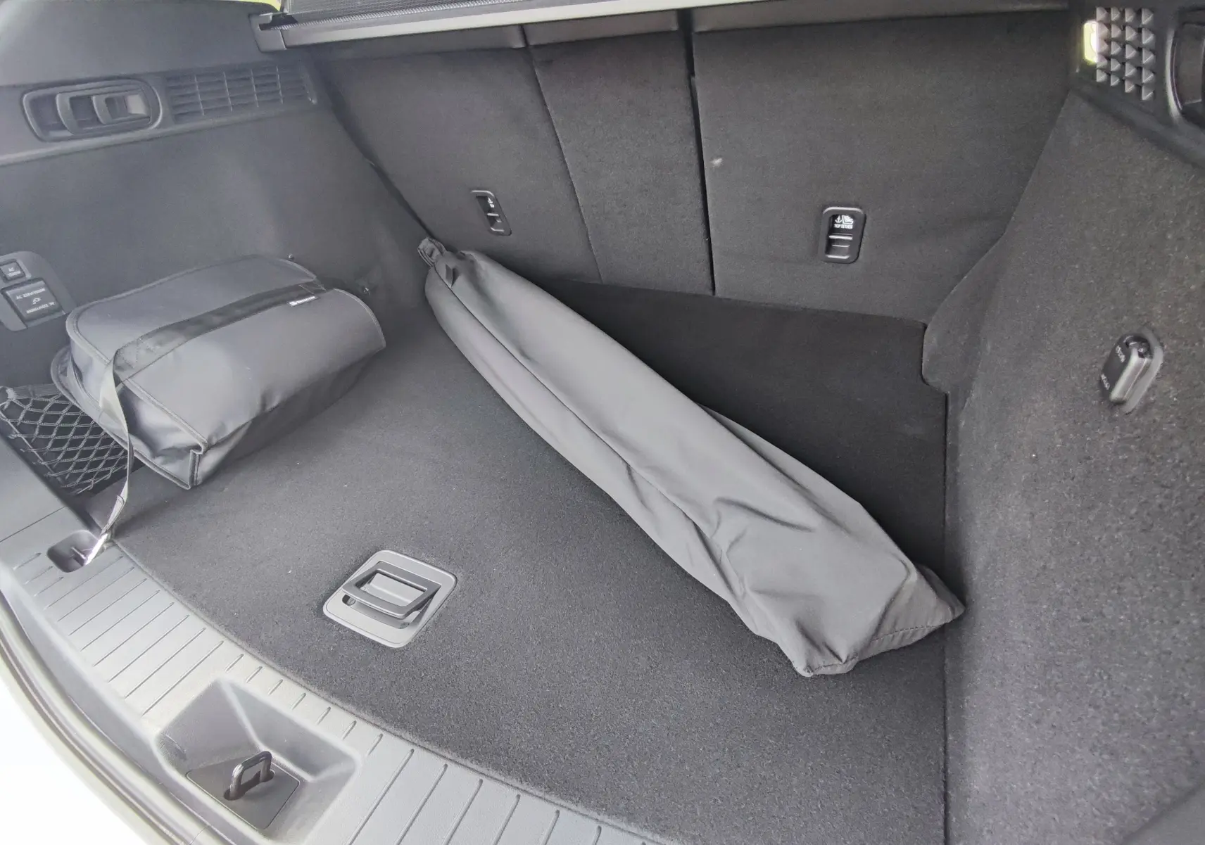 Coffre ouvert du Mazda CX-60 blanc métal 2024, vue intérieure montrant un rangement noir et un sac plié sur le tapis.