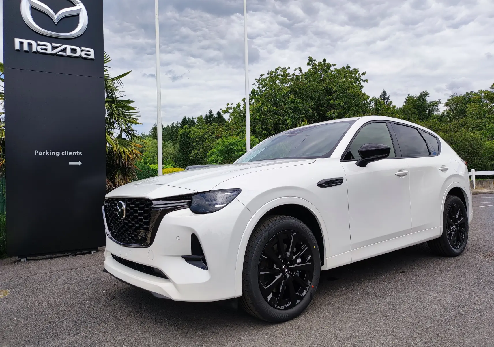 Vue 3/4 avant droit d'un Mazda CX-60 blanc métal avec jantes noires, devant un panneau Mazda en extérieur.