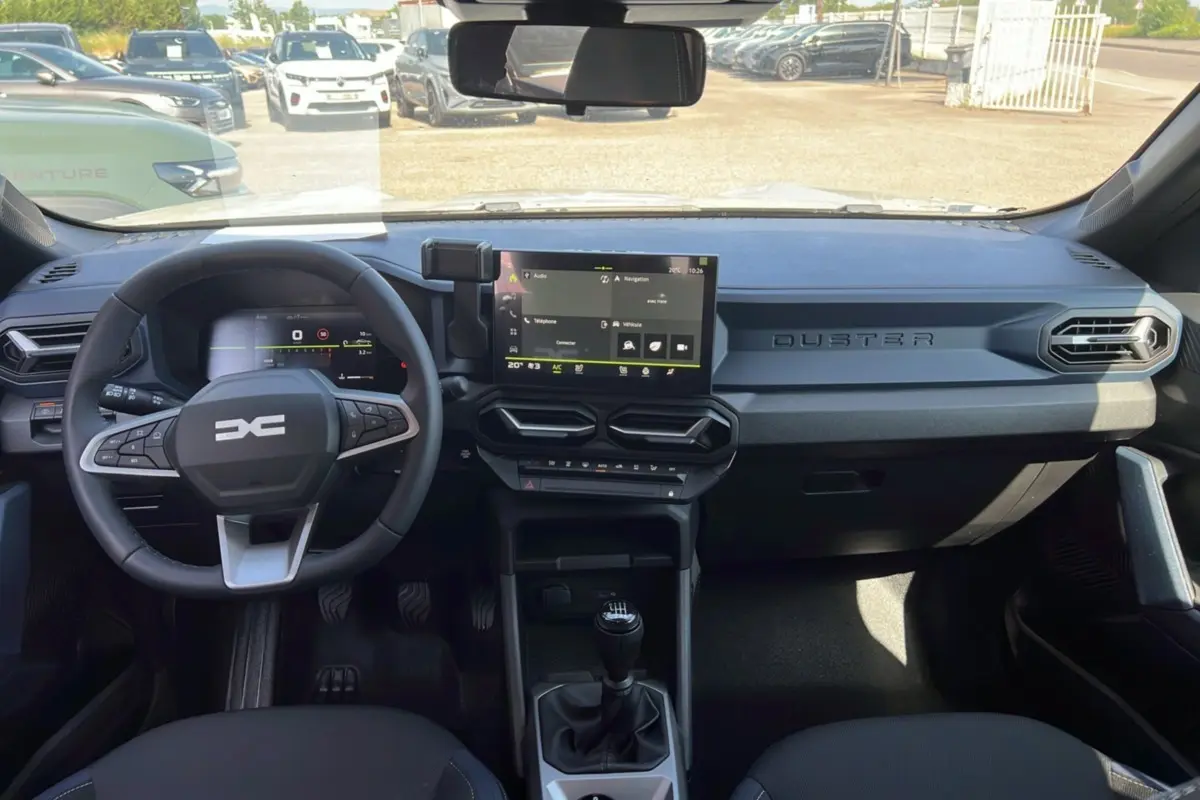 Vue intérieure du tableau de bord du Dacia Duster 2025 avec volant multifonction, écran tactile central et levier de vitesses manuel.