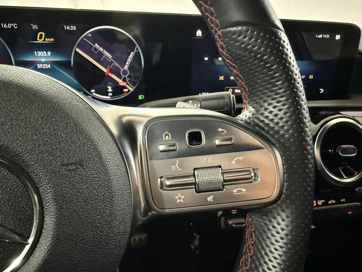 Gros plan sur le volant cuir noir avec surpiqûres rouges et commandes multifonctions d'une Mercedes Classe A gris foncé, tableau de bord digital visible.