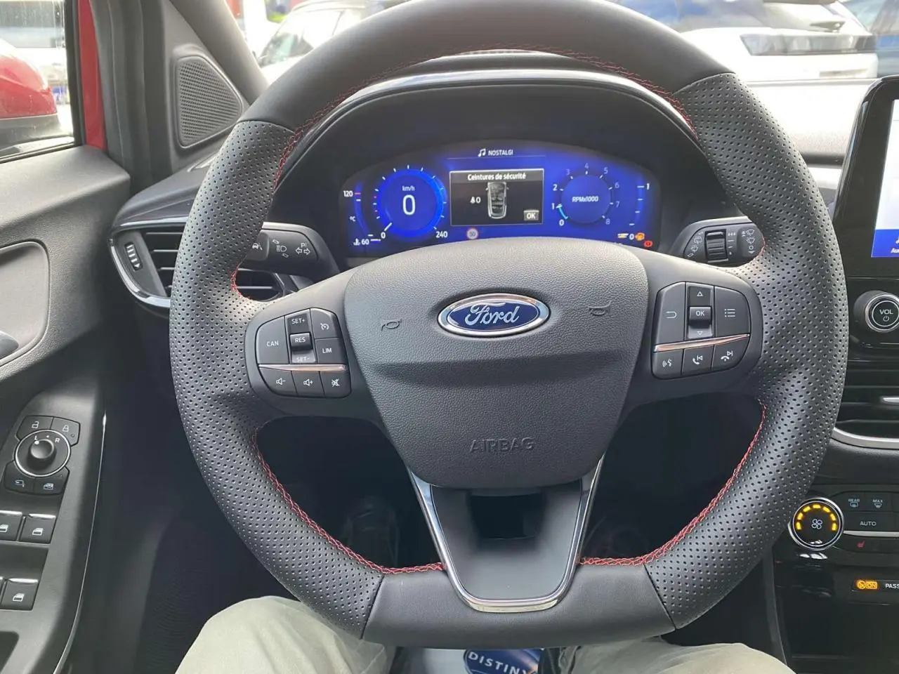 Vue intérieure centrée sur le volant perforé avec surpiqûres rouges du Ford Puma 2022, tableau de bord numérique allumé.
