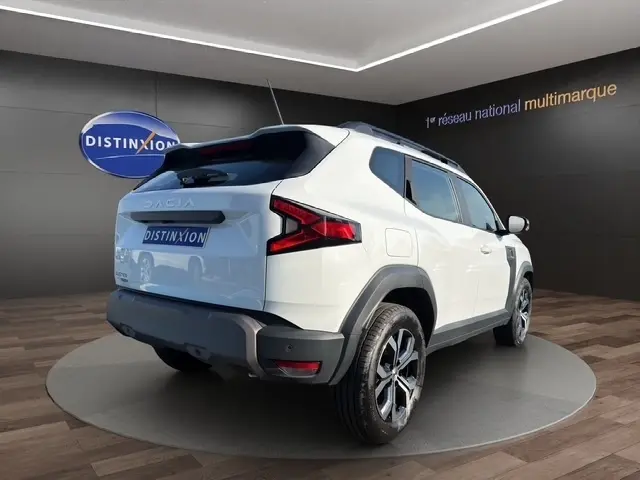 Vue 3/4 arrière droite d'un Dacia Duster blanc 2025 avec jantes alliage et feux arrière LED distinctifs dans un showroom.