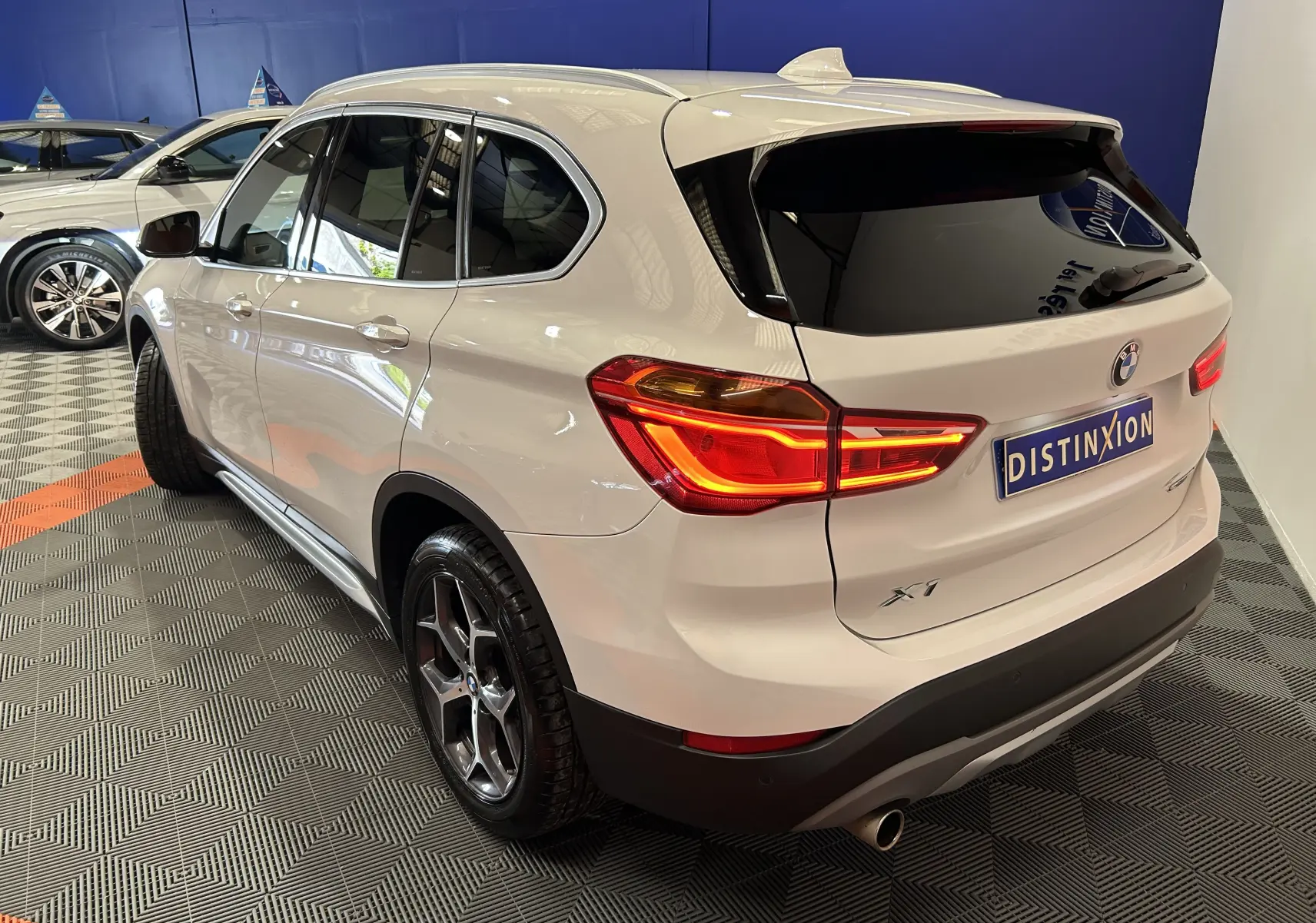 Vue 3/4 arrière droite d'un BMW X1 blanc avec feux allumés et hayon électrique en intérieur showroom.