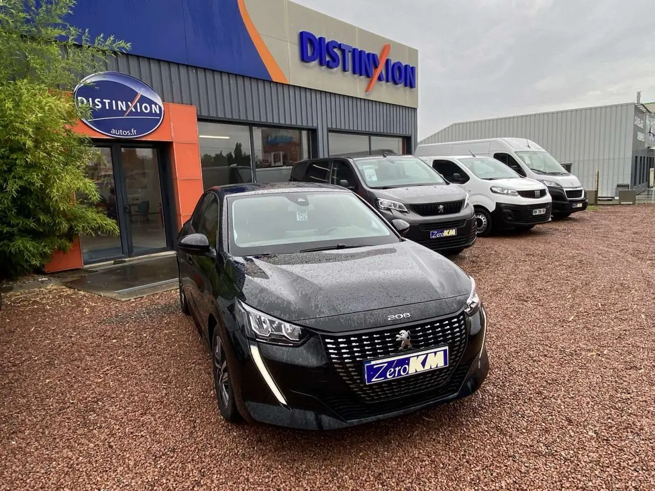 Peugeot 208 noir métallisé vue 3/4 avant sur parking devant concession Distinxion par temps pluvieux.