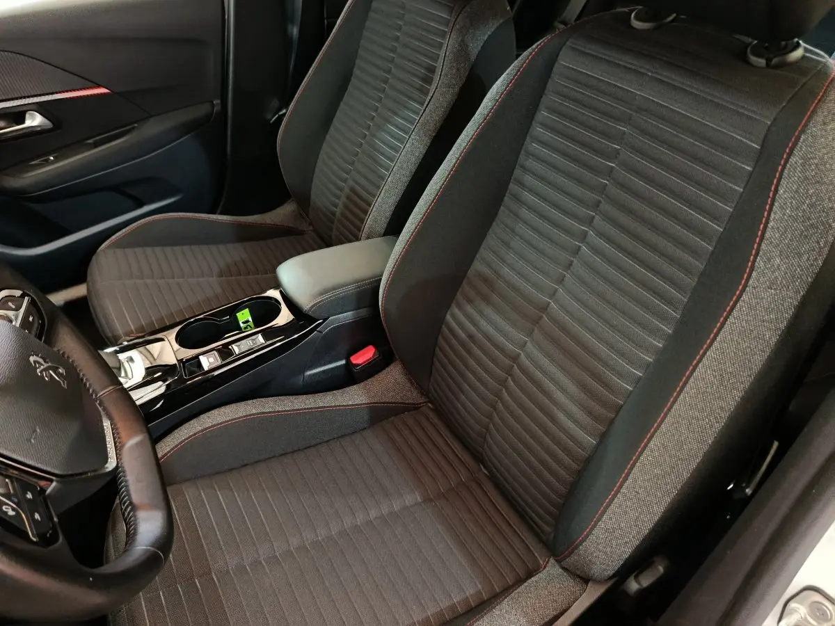 Vue intérieure rapprochée des sièges avant noirs avec surpiqûres rouges et console centrale d’une Peugeot 208 Electrique blanche.